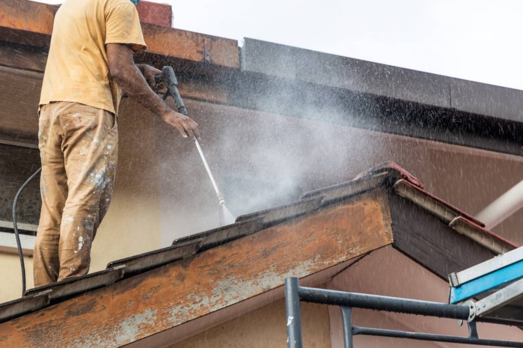 Worker using high pressure water jet spray gun to wash and clean dirt from rooftop tile at residential building in renovation